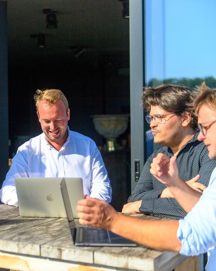 ¿Apoyo para una mayor eficiencia en Odoo? dos empleados de la empresa de servicios acompañados por el fundador Arnaud Percy alrededor de una mesa de reuniones al aire libre con dos ordenadores portátiles mac windows ios tres hombres en camisa sin corbata con camisas de diferentes colores
